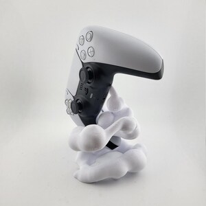 Op de afbeelding: Een witte en zwarte PlayStation 5-controller op een witte, wolkvormige standaard. De controller heeft zwarte knoppen en een zwart touchpad. De standaard is een decoratief accessoire.