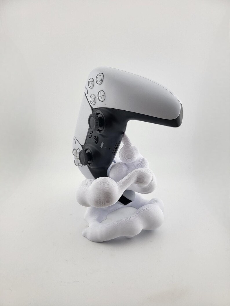Op de afbeelding: Een witte en zwarte PlayStation 5-controller op een witte, wolkvormige standaard. De controller heeft zwarte knoppen en een zwart touchpad. De standaard is een decoratief accessoire.