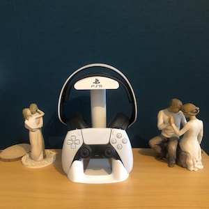 May include: White PS5 controller charging station with a black and white headset. The station has the PS5 logo and button symbols. Two small figurines are on either side of the station. The background is a dark teal color.