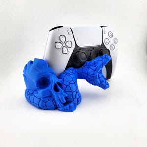 Könnte beinhalten: Ein weiß-schwarzer Gamecontroller befindet sich hinter einer blauen Schädel- und Schlangenskulptur. Die blaue Figur hat ein strukturiertes, rissiges Muster. Der Controller hat ein Touchpad und verschiedene Tasten.