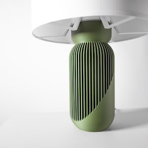 Puede incluir: Una l&aacute;mpara de mesa verde moderna con una pantalla blanca. La base de la l&aacute;mpara tiene un dise&ntilde;o cil&iacute;ndrico con ranuras verticales y un recorte diagonal. La pantalla tiene forma circular plana.