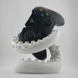 May include: A black Xbox controller rests in a white octopus-shaped stand. The controller has colored buttons and a directional pad. The stand is a decorative item for gamers.