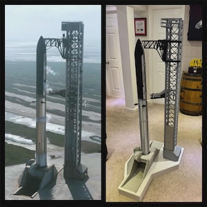 May include: A scale model of a silver and black rocket standing next to a tall gray launch tower structure on a white base. The rocket features a pointed nose cone and a cylindrical body, positioned beside a detailed lattice-style support tower.