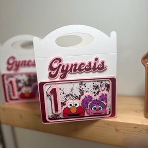 May include: White party favor box with a handle, featuring the name "Gynesis" in pink glitter lettering. The box has a clear window displaying Elmo and Abby Cadabby from Sesame Street, along with the number "1".