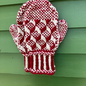 May include: A red and white knitted mitten with a geometric pattern. The mitten is hanging against a green background. The cuff features red and white stripes.