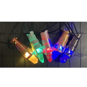 May include: Five glowing pendant lights in various colors: amber, green, red, blue, and purple. Each light has a metallic casing and a clear, pointed end. The pendants are strung on black cords.