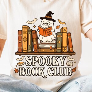 May include: A cream-coloured t-shirt featuring a graphic of a ghost wearing a witch hat and glasses, reading a book while sitting on a stack of books on a shelf. The text below reads SPOOKY BOOK CLUB, accompanied by small bat and leaf illustrations.