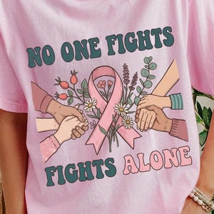 以下が含まれることがあります： 淡いピンク色の半袖Tシャツ。ピンク色のリボンと花の周りで複数の手が繋ぎ合っているグラフィックが描かれています。上部にはダークグリーンの文字で「NO ONE FIGHTS」、下部にはピンク色の文字で「FIGHTS ALONE」とプリントされています。