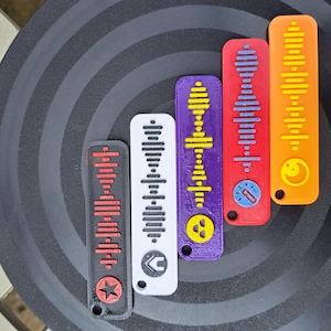 May include: Six colourful keychains with a sound wave design. Each keychain has a different colour and a small symbol at the bottom. The colours are red, white, purple, orange, and black.