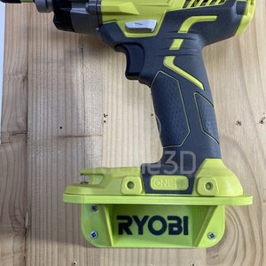 Peut inclure: Une perceuse Ryobi jaune et noire avec une poignée noire et un support mural jaune et noir. Le support mural porte le logo Ryobi.