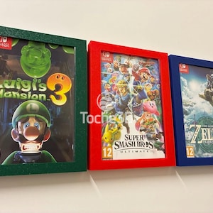 Peut inclure: Trois couvertures de jeux Nintendo Switch encadrées : "Luigi's Mansion 3" dans un cadre vert, "Super Smash Bros. Ultimate" dans un cadre rouge et "The Legend of Zelda: Tears of the Kingdom" dans un cadre bleu. Chaque cadre a une finition texturée.