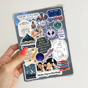 May include: A clear case covered in stickers, including text that reads "THERE YOU ARE. I've been looking for you" and "Hello, Feyre Darling." The stickers feature illustrations of mountains, books, and other designs. The case is held by a hand with red nail polish.