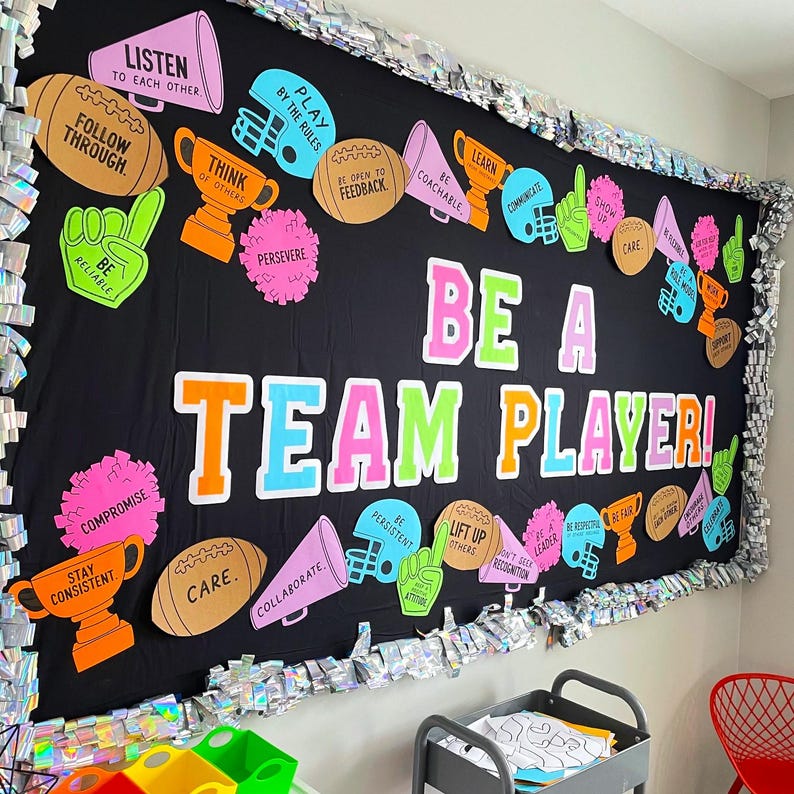 May include: A black bulletin board display featuring the text BE A TEAM PLAYER in large colorful letters. The board is decorated with cutouts of footballs, trophies, megaphones, and foam fingers, each printed with motivational phrases.