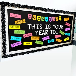 May include: A rectangular notice board with a black background and a white wood-grain frame featuring a black and white polka dot border. The board displays the text 2026-2027, THIS IS YOUR YEAR TO..., and various colourful notes with motivational phrases.