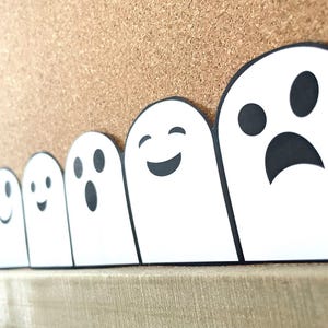 May include: A row of white ghost cutouts with black facial expressions, including smiles and a sad face. The ghosts are arranged in a line against a corkboard background, creating a Halloween or spooky theme. The cutouts are on a wooden surface.