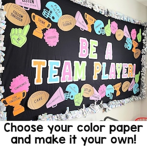 May include: A black bulletin board display featuring the text BE A TEAM PLAYER in large colorful letters. The board is decorated with cutouts of footballs, trophies, megaphones, foam fingers, and pom-poms, each printed with motivational phrases.