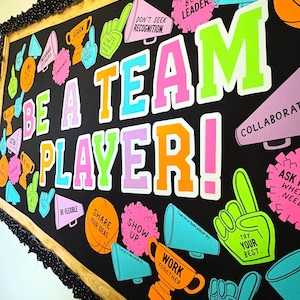 May include: A black bulletin board display featuring the text BE A TEAM PLAYER! in large, colorful block letters. The board is decorated with cutouts of megaphones, trophies, foam fingers, and pom-poms in neon pink, orange, blue, and green.