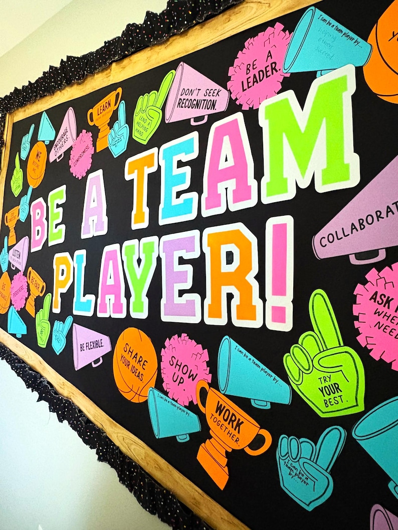 May include: A black bulletin board display featuring the text BE A TEAM PLAYER! in large, colorful block letters. The board is decorated with cutouts of megaphones, trophies, foam fingers, and pom-poms in neon pink, orange, blue, and green.
