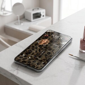 Puede incluir: Un smartphone muestra un sitio web con fondo de estampado de leopardo, el texto "NAILS by Aliyah" y un retrato circular. Junto al tel&eacute;fono hay un frasco de esmalte de u&ntilde;as rosa empolvado y un pincel fino para u&ntilde;as sobre una superficie blanca.
