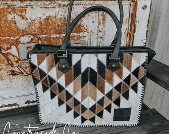 Puede incluir: Bolso tote con estampado geométrico con asas y ribetes de cuero negro. El bolso presenta un diseño de patchwork con formas triangulares blancas, negras y marrones. El bolso tiene forma rectangular y un borde cosido. Las palabras "Countryside Co." están escritas abajo a la izquierda.