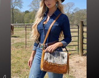 Puede incluir: Una mujer con camisa y vaqueros de mezclilla, sombrero de vaquero negro y pendientes turquesa. Lleva un bolso bandolera de cuero marrón con frente de piel de vaca y detalles decorativos.