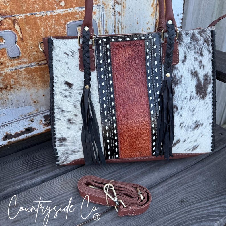 May include: A cowhide leather tote bag with a brown leather shoulder strap. The bag features a central panel of textured brown leather, flanked by panels of black and white cowhide. Braided leather accents and tassels add detail.