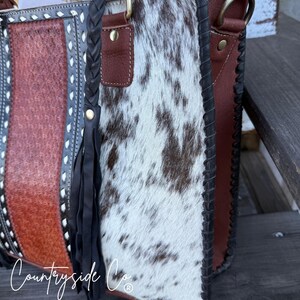 May include: A cowhide leather handbag with a brown, white, and black spotted pattern. The bag features a braided handle, leather accents, and a decorative tassel. The bag has a brown and white spotted pattern.