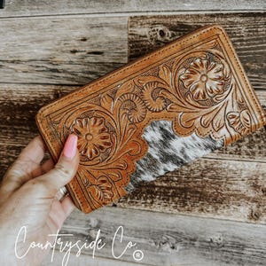 May include: A brown leather wallet with a floral tooled design and a white and black cowhide patch. The wallet has a zipper closure and is held in a hand.