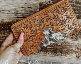 May include: A brown leather wallet with a floral tooled design and a white and black cowhide patch. The wallet has a zipper closure and is held in a hand.