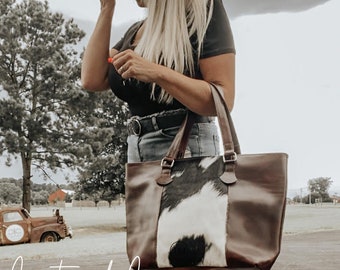 May include: A brown leather tote bag with a black and white cowhide panel. The bag has two top handles and a zipper closure.