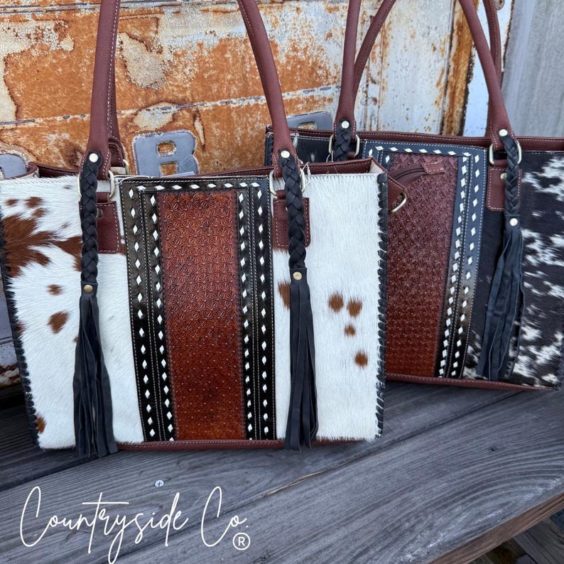 May include: Three cowhide tote bags with brown leather handles and accents. The bags feature a mix of white, brown, and black cowhide patterns, with a central embossed leather panel. Each bag has a braided handle detail and a black tassel.