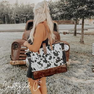 May include: A large tote bag with a cowhide pattern in black and white, complemented by brown leather accents. The bag features a decorative embossed leather base and leather straps. The Countryside Co. logo is visible in the bottom left corner.
