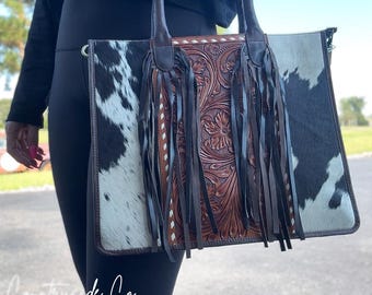 May include: A rectangular tote bag with a cowhide pattern on the sides and a brown, floral-tooled leather center. The bag has long brown handles and dark brown fringe. The text "Countryside Co." is visible at the bottom.