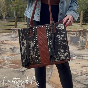 May include: A large, rectangular handbag with a cowhide pattern in black and white. The center panel is brown with white stitching. The bag has brown leather handles and black tassels. The text "Countryside Co." is visible at the bottom.