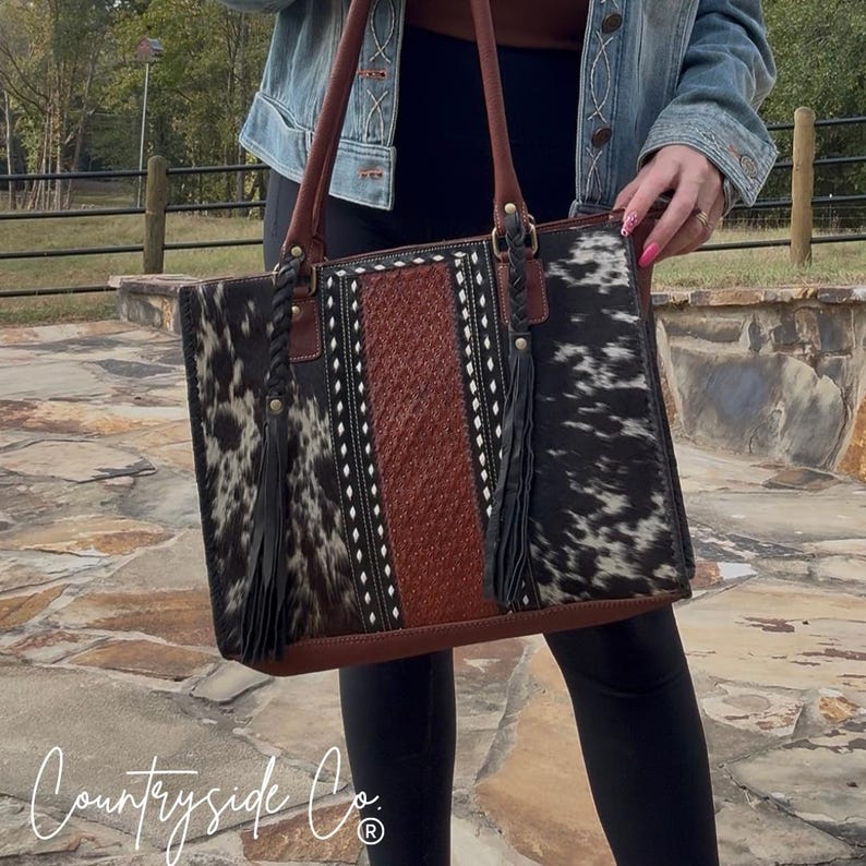 May include: A large, rectangular handbag with a cowhide pattern in black and white. The center panel is brown with white stitching. The bag has brown leather handles and black tassels. The text "Countryside Co." is visible at the bottom.
