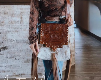 May include: A brown leather crossbody bag with a tooled design and fringe accents. The bag has a white cowhide flap and a silver buckle. The bag is worn by a person wearing a brown patterned shirt and jeans. The text "Countryside Co." is visible in the bottom left corner of the image.
