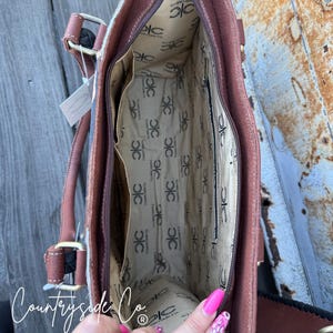 May include: Open view of a brown leather handbag with a patterned interior and multiple pockets. The bag has brown handles and a zipper closure. The interior lining features a repeating logo pattern. The brand name "Countryside Co." is visible.