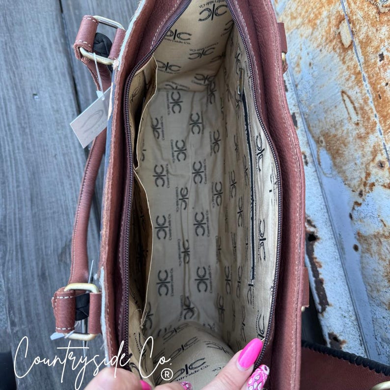 May include: Open view of a brown leather handbag with a patterned interior and multiple pockets. The bag has brown handles and a zipper closure. The interior lining features a repeating logo pattern. The brand name "Countryside Co." is visible.