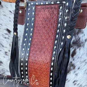 May include: A close-up of a handcrafted leather tote bag. The bag features a central panel with a floral embossed pattern in shades of brown. The edges are trimmed with black leather, white stitching, and diamond-shaped accents. Braided black leather handles and fringe complete the design.