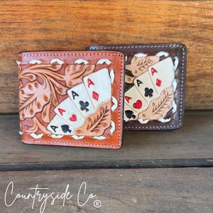 May include: Two leather wallets with embossed playing card designs. One wallet is light brown with a floral pattern and the other is dark brown. Both wallets feature the Ace of Spades, Hearts, Diamonds, and Clubs.