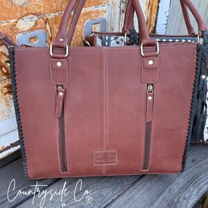 May include: A brown leather tote bag with two zippered pockets and a stitched design. The bag has two handles and a decorative black braid on the side. The text "Countryside Co." is visible at the bottom.