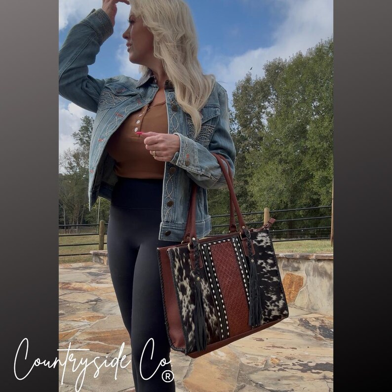 May include: A brown and black cowhide tote bag with a leather handle and a woven center panel. The bag has black fringe and the text "Countryside Co" in a cursive font.