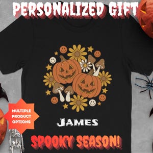 Könnte beinhalten: Ein schwarzes T-Shirt mit einer Grafik aus zwei Kürbissen, Pilzen, Blumen, Sternen und Smileys. Der Name JAMES ist in Weiß unter der Grafik gedruckt, mit dem Text SPOOKY SEASON! in Orange und Rot am unteren Rand des T-Shirts.