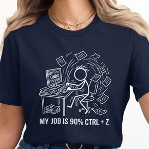 Può includere: T-shirt blu navy con grafica bianca di un omino stilizzato al computer, circondato da fogli volanti. Lo schermo del computer mostra "UNDO". Il testo "MY JOB IS 90% CTRL + Z" è sotto l'immagine.