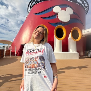 Może przedstawiać: Biała koszulka z napisem "SEIZE THE MOMENT" i "LET YOUR FANTASIES AND DREAMS SET SAIL" na grafice postaci Disneya. Tło przedstawia czerwoną i białą konstrukcję z uszami Myszki Miki.