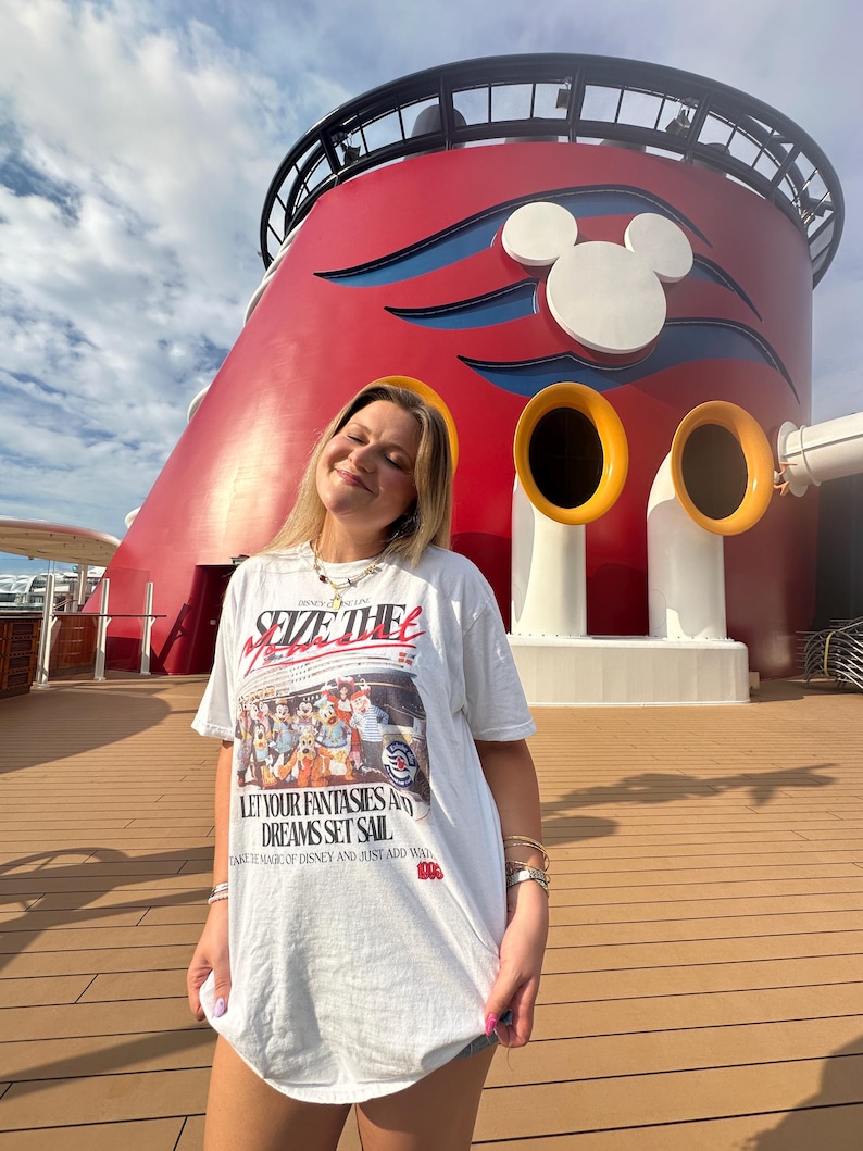 Może przedstawiać: Biała koszulka z napisem "SEIZE THE MOMENT" i "LET YOUR FANTASIES AND DREAMS SET SAIL" na grafice postaci Disneya. Tło przedstawia czerwoną i białą konstrukcję z uszami Myszki Miki.