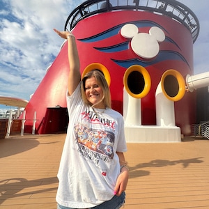 Może przedstawiać: Biały t-shirt z nadrukiem graficznym przedstawiającym postacie z kresk&oacute;wek. Koszulka jest połączona z dżinsowymi szortami. Tło obejmuje czerwoną i białą konstrukcję z uszami Myszki Miki i ż&oacute;łtymi akcentami na tle błękitnego nieba.