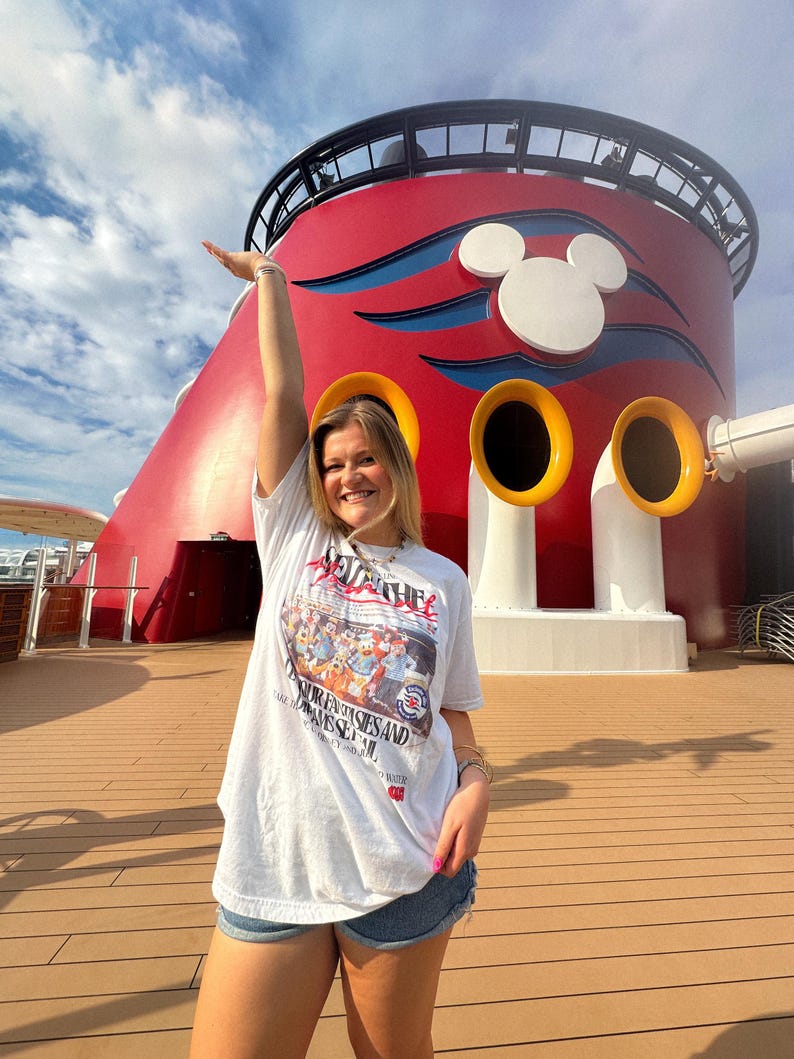 Może przedstawiać: Biały t-shirt z nadrukiem graficznym przedstawiającym postacie z kresk&oacute;wek. Koszulka jest połączona z dżinsowymi szortami. Tło obejmuje czerwoną i białą konstrukcję z uszami Myszki Miki i ż&oacute;łtymi akcentami na tle błękitnego nieba.