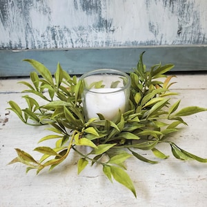 May include: A green leafy wreath with a clear glass votive candle holder in the center. The candle holder is filled with a white candle.