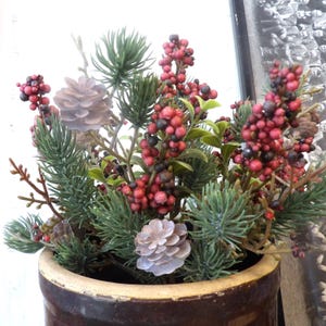 May include: A decorative arrangement of faux greenery and berries in a brown ceramic pot. The arrangement includes pine needles, red berries, and pinecones. The pot has a wide, slightly flared rim.